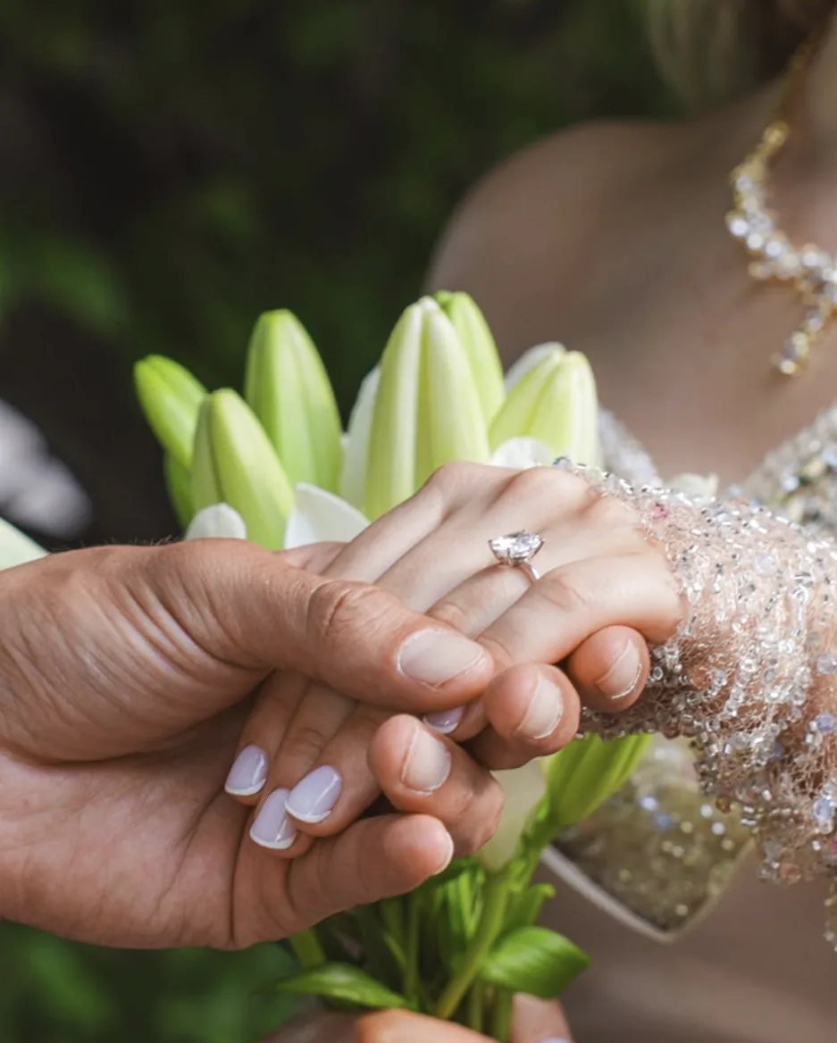 Bräutigam hält die Hand der Braut mit Verlobungsring, daneben weiße Lilien
